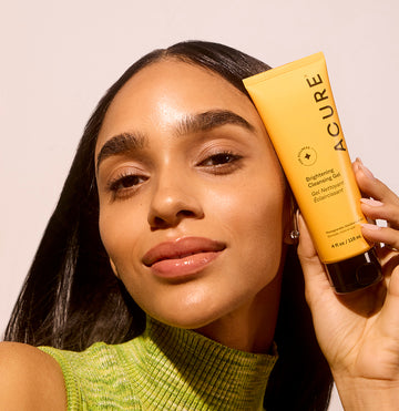 Woman holding a tube of Acure Brightening Cleansing gel next to her glowing face skin
